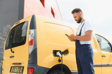 Kurye, yoğun iş günlerinde elektrikli minibüsü şarj ederken akıllı telefonu kontrol ediyor, modern teknolojiyi ve sürdürülebilir ulaşımı sergiliyor