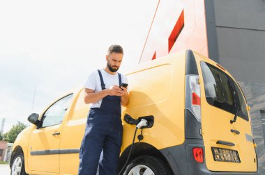 Kurye, sarı elektrikli minibüsünün şarj olmasını beklerken akıllı telefonunu kontrol ediyor. Modern teknolojiyi sürdürülebilir ulaşım aracıyla harmanlıyor.