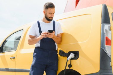 Kurye şarj istasyonunda elektrik dağıtım aracını şarj ederken akıllı telefonunu kontrol ediyor.