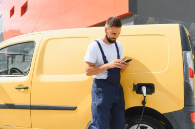 Şehir merkezinde elektrikli minibüsü şarj ederken akıllı telefon kullanan kurye, teknolojiyi sürdürülebilir ulaşım ile harmanlıyor