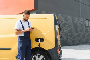 Elektrik dağıtım minibüsü, sürdürülebilir ulaşım ve dağıtım hizmeti şarj ederken akıllı telefon kullanan kargo işçisi