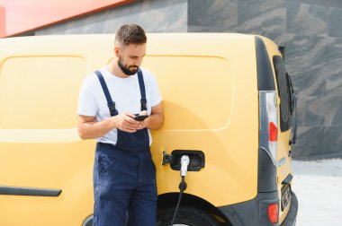 Kurye şarj istasyonunda elektrik dağıtım aracını şarj ederken akıllı telefonunu kontrol ediyor.