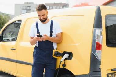 Şehir merkezinde elektrikli minibüsü şarj ederken akıllı telefon kullanan kargo şoförü, teknolojiyi dengelemek ve sürdürülebilir ulaşım