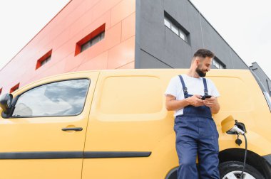 Sarı elektrikli minibüsü modern bir binanın önünde şarj olurken işçi akıllı telefonunu kullanıyor.