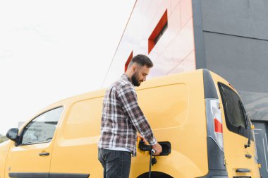 Şehir ortamında modern bir binanın yakınındaki bir şarj istasyonunda elektrik dağıtım minibüsü şarj eden bir teslimat şoförü.