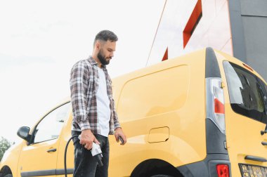 Yükleme kablosu tutan kargo şoförü ve şarj istasyonunda sarı elektrikli minibüsün şarj portu açılıyor.