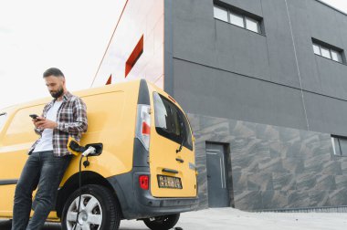 Elektrikli minibüsü modern binanın önünde şarj olurken teslimatçı akıllı telefon kullanıyor.