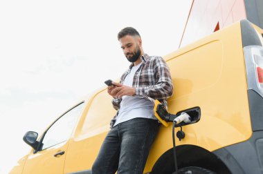 Sarı elektrikli minibüsü şarj istasyonunda şarj olurken akıllı telefon kullanan teslimatçı.