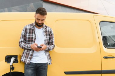 Elektrikli minibüsü şarj olurken akıllı telefon kullanan teslimat şoförü, modern dağıtım hizmetlerini ve sürdürülebilir ulaşım hizmetlerini temsil ediyor.