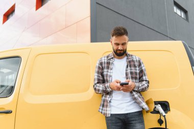 Elektrikli minibüsü modern binanın önünde şarj olurken teslimatçı akıllı telefon kullanıyor.