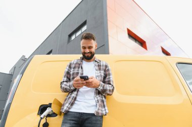 Elektrikli minibüsü modern binanın önünde şarj olurken teslimatçı akıllı telefon kullanıyor.