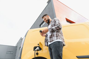 Elektrikli minibüsü modern binanın önünde şarj olurken teslimatçı akıllı telefon kullanıyor.