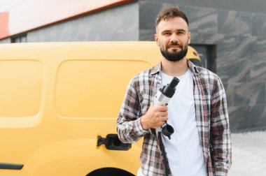 Sarı minibüsünün yanında elektrikli araç şarjı tutan teslimatçı, sürdürülebilir ulaşım ve çevre dostu dağıtım hizmetlerini destekliyor.