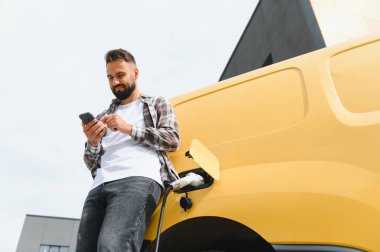 Şehir şarj istasyonunda elektrikli minibüsü şarj ederken akıllı telefon kullanan teslimat sürücüsü, sürdürülebilir şehir ulaşımını kucaklıyor