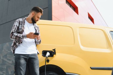 Elektrikli minibüsü modern binanın önünde şarj olurken teslimatçı akıllı telefon kullanıyor.