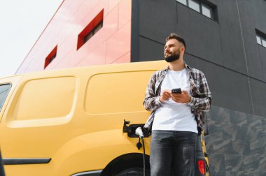 Elektrikli minibüsü modern bir binanın önünde şarj olurken akıllı telefon kullanan teslimatçı.