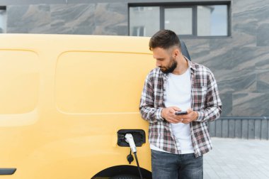 Şehir merkezinde elektrikli minibüsü şarj ederken akıllı telefon kullanan teslimat sürücüsü, modern teknoloji ve sürdürülebilirliği kucaklıyor