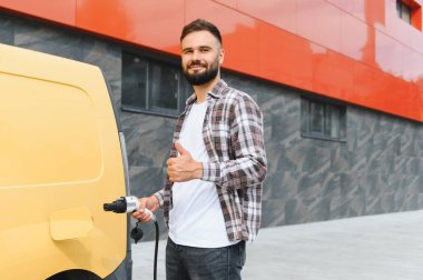 Şarj kablosu tutan teslimatçı, elektrik dağıtım aracının yanında, sürdürülebilir ve çevre dostu taşımacılığın simgesi olarak baş parmak işareti yapıyor.