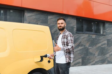 Nakliye şöförü elektrikli minibüsü şarj ediyor ve modern bir binanın önünde baş parmak kaldırıyor. Sürdürülebilir taşımacılığa bağlılığını gösteriyor.