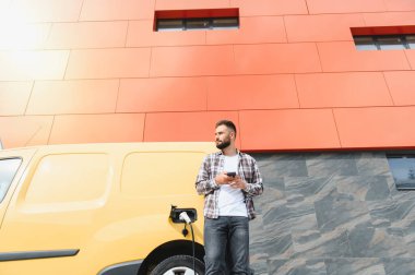 Elektrikli minibüsü modern binanın önünde şarj olurken teslimatçı akıllı telefon kullanıyor.