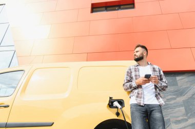 Elektrikli minibüsü modern binanın önünde şarj olurken teslimatçı akıllı telefon kullanıyor.