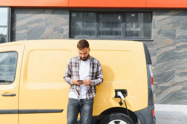 Elektrikli minibüsü modern binanın önünde şarj olurken teslimatçı akıllı telefon kullanıyor.