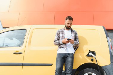 Elektrikli minibüsü renkli bir binanın önünde şarj olurken teslimatçı akıllı telefon kullanıyor.