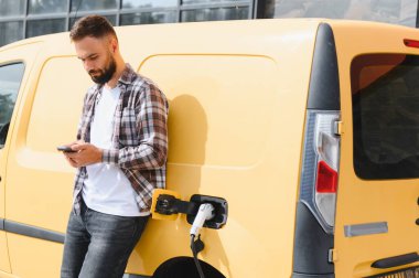 Şehir merkezinde elektrikli bir minibüsü şarj ederken akıllı telefon kullanan teslimat sürücüsü, modern ulaşım ve sürdürülebilir enerjiyi vurguluyor