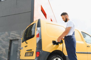 Nakliye şöförü rotayı tamamladıktan sonra elektrikli minibüsü şarj ediyor, modern dağıtım hizmetlerinde sürdürülebilir lojistik ve çevre dostu ulaşımı sembolize ediyor