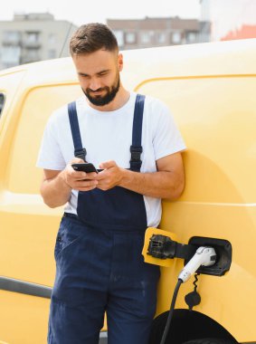 Kurye akıllı telefonunu kontrol ederken elektrikli minibüsü şarj ediyor, teknoloji ve kentsel taşımacılıkta sürdürülebilirliği birleştiriyor