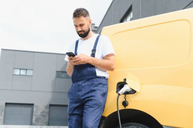 Teslimatçı akıllı telefonu kontrol ederken bir depoda elektrikli minibüsü şarj ediyor, modern teknolojiyi ve sürdürülebilir ulaşımı vurguluyor.