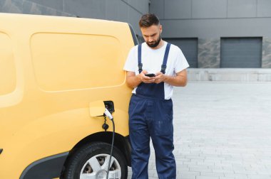 Kurye akıllı telefon kullanırken sarı elektrikli minibüsü modern bir binanın önünde şarj oluyor.