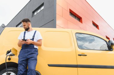 Kurye, modern bir binanın yanında elektrik dağıtım kamyonunu şarj ederken akıllı telefonunu kontrol ediyor ve sürdürülebilir ulaşım çözümlerini kucaklıyor.