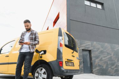 Elektrikli minibüsü modern binanın önünde şarj olurken teslimatçı akıllı telefon kullanıyor.