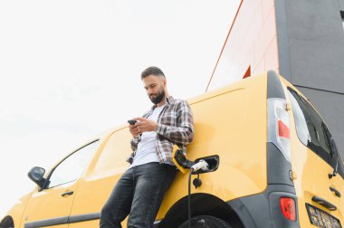 Elektrikli minibüsü modern binanın önünde şarj olurken teslimatçı akıllı telefon kullanıyor.