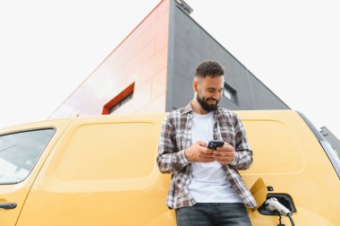 Şık, modern bir binanın önünde elektrikli minibüsü şarj ederken akıllı telefonu kontrol eden teslimatçı, harmanlama teknolojisi ve sürdürülebilirlik