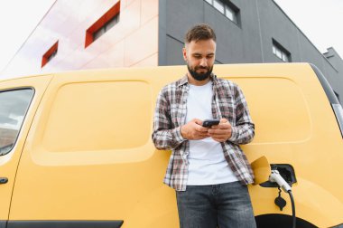 Elektrikli minibüsü modern bir binanın önünde şarj olurken akıllı telefon kullanan teslimatçı.