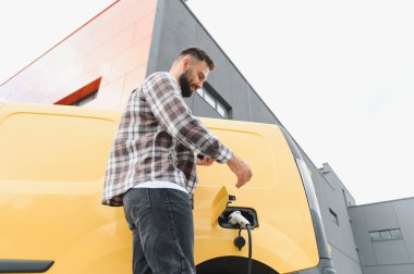 Bir şarj istasyonunda elektrikli minibüsü şarj ederken, sürdürülebilir ulaşımı kucaklayan bir cep telefonu uygulamasına bağlanan teslimat sürücüsü