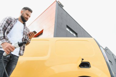 Teslimat sürücüsü kabloyu tutuyor ve elektrik dağıtım minibüsünü şarj ederken akıllı telefon uygulaması kullanıyor.