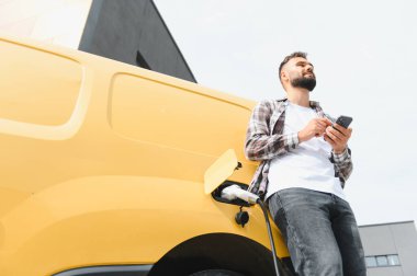 Güneşli bir şehir gününde şık, modern bir binanın önünde elektrikli minibüs şarj ederken akıllı telefon kullanan teslimatçı.