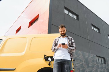 Elektrikli minibüsü modern binanın önünde şarj olurken teslimatçı akıllı telefon kullanıyor.