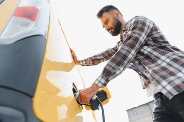 Halka açık bir şarj istasyonunda şarj kablosunu sarı elektrikli araca bağlayan genç yetişkin, sürdürülebilir ulaşımı teşvik ediyor