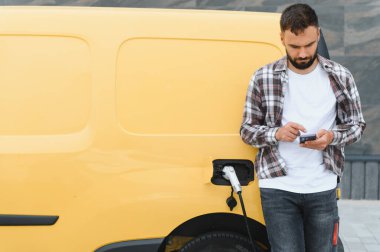 Bir istasyonda elektrikli minibüsü şarj ederken akıllı telefonu kontrol eden teslimatçı, modern teknolojiyi sürdürülebilir ulaşım ile harmanlıyor