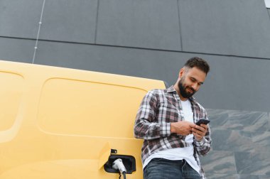 Teslimatçı, modern bir binanın önünde elektrikli bir minibüsü şarj ederken akıllı telefon kullanıyor, teknolojiyi sürdürülebilir ulaşım çözümleriyle harmanlıyor.