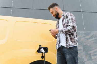 Teslimatçı, kentsel bir ortamda elektrik dağıtım kamyonunu şarj ederken akıllı telefonu kontrol ediyor, sürdürülebilir ulaşımı vurguluyor