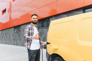 Elektrik kablosu tutan teslimatçı modern binanın yanındaki şarj istasyonunda elektrik dağıtım kamyonunu şarj ederken baş parmağını kaldırıyor.