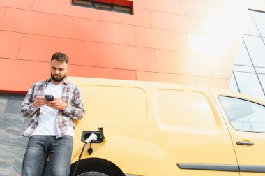 Elektrikli minibüsü modern binanın önünde şarj olurken teslimatçı akıllı telefon kullanıyor.