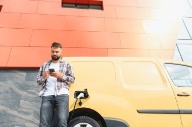 Elektrikli minibüsü modern binanın önünde şarj olurken teslimatçı akıllı telefon kullanıyor.