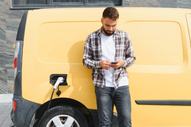 Bir şarj istasyonunda elektrikli minibüsü şarj ederken akıllı telefonu kontrol eden teslimatçı modern teknoloji ve verimliliği kucaklıyor.