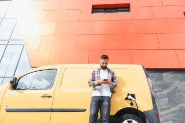 Elektrikli minibüsü modern binanın önünde şarj olurken teslimatçı akıllı telefon kullanıyor.